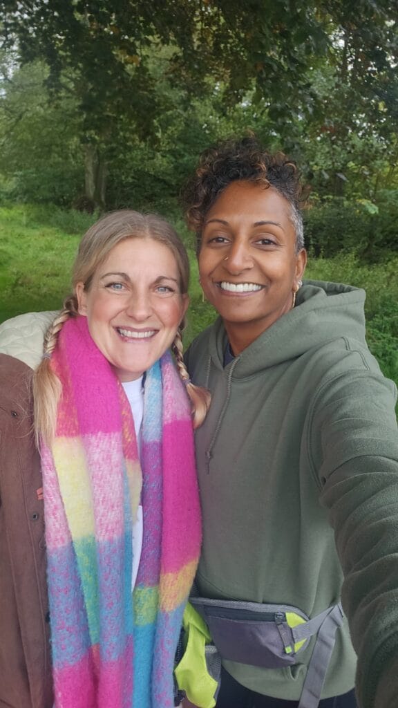 Two people are standing close together outdoors, smiling warmly at the camera. The person on the left has light hair styled in two braids and is wearing a colourful scarf in shades of pink, yellow and blue along with a brown jacket. The person on the right has short curly hair and is dressed in a green hoodie with a small grey and yellow waist bag. They are surrounded by lush green trees and grass, creating a peaceful woodland backdrop.