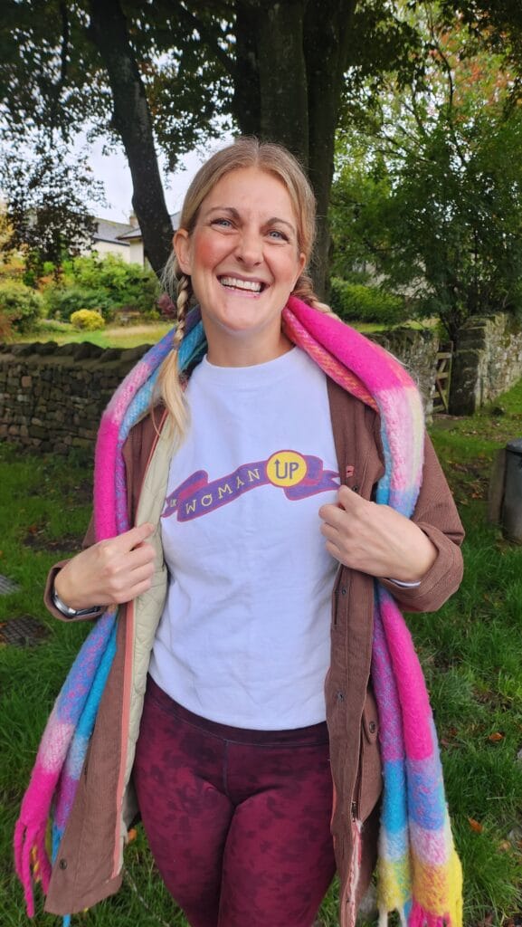 A woman stands outdoors smiling brightly, holding the sides of her open brown jacket to show a white T shirt with a colourful “Woman Up” design. She has light hair in two braids and is wrapped in a vibrant pink, blue and yellow scarf. Behind her is a stone wall, green grass and leafy trees, giving the scene a relaxed, natural feel.
