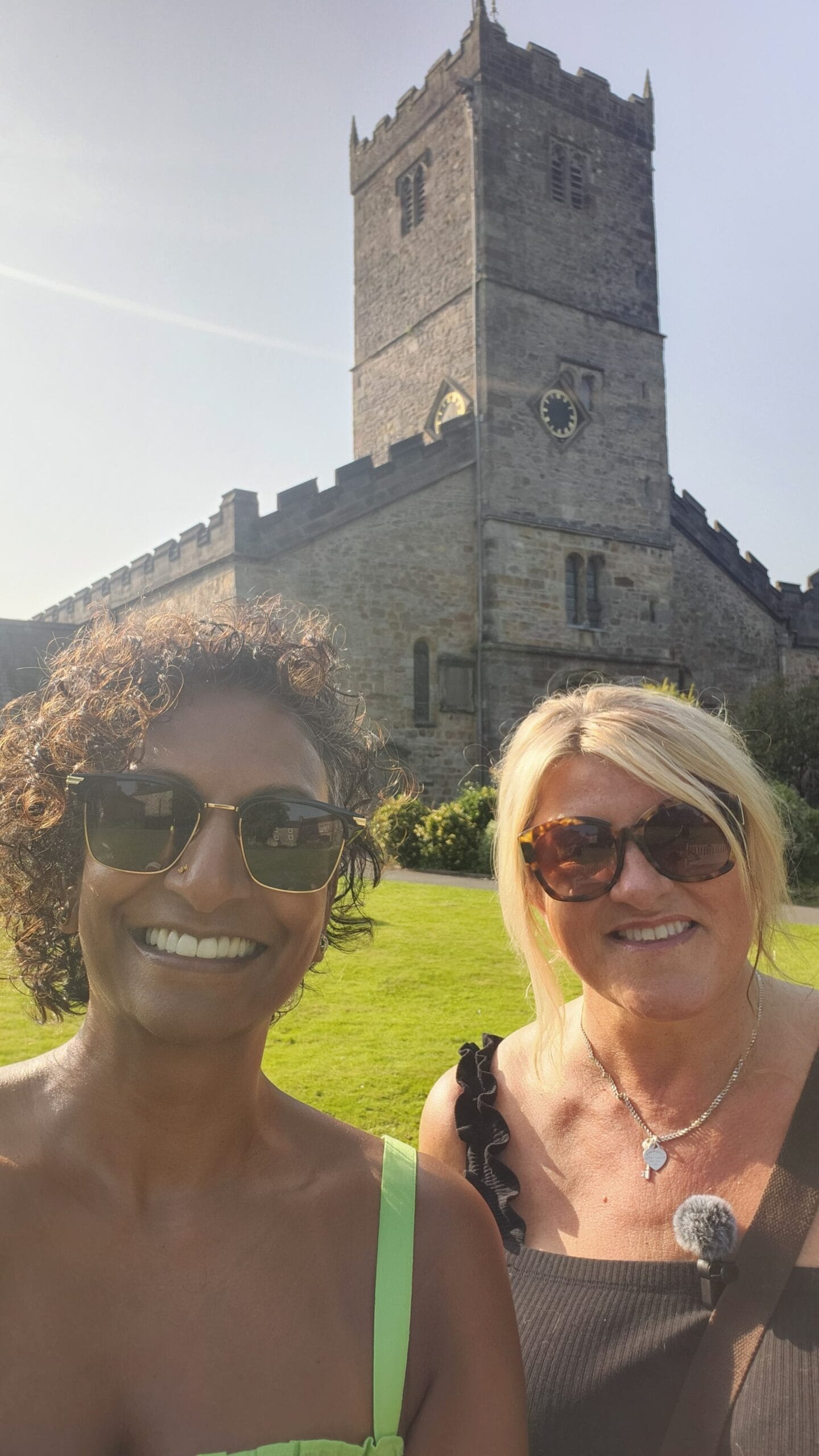 Two women smiling in bright sunlight with a large stone church behind them. The church has a tall square tower, battlements along the roofline and a clock face. They are standing on a green lawn, both wearing sunglasses.