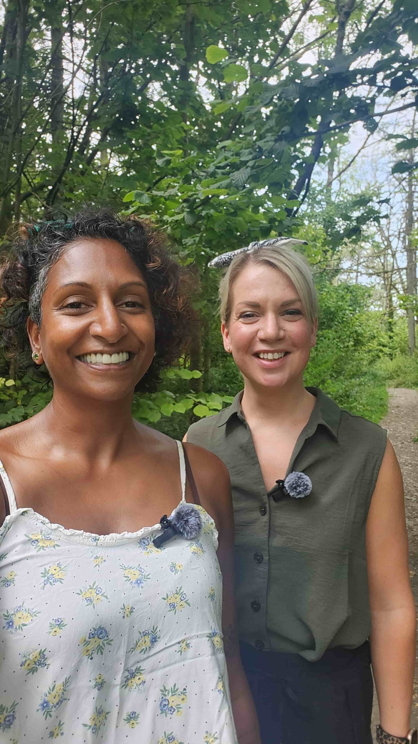 Two women smiling at the camera while walking along a woodland path, both wearing clip on microphones, surrounded by green trees and summer foliage.